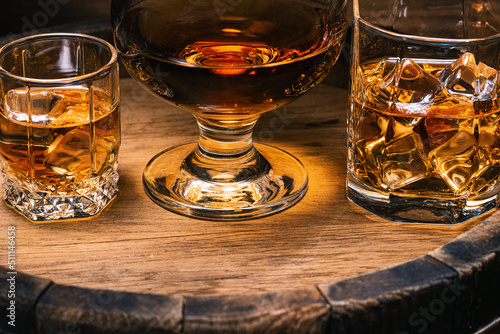 Alcoholic beverages whiskey, cognac, brandy, stand in glasses with ice on wooden barrel of whiskey close-up. Drink whiskey or cognac with ice in bar and spilled drinks directly from barrel.