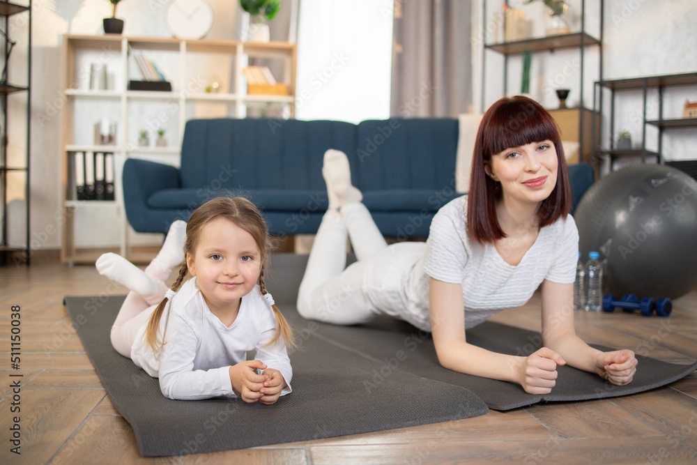 A young caucasian mother and her little daughter lying on a mat on the floor during fitness classes in the living room at home and smiling at the camera. Healthy Lifestyle A young caucasian mother and her little daughter lying on a mat on the floor during fitness classes in the living room at home and smiling at the camera. Healthy Lifestyle