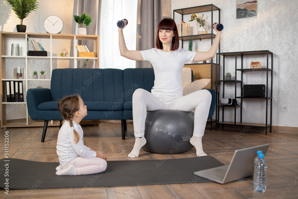 Positive young woman doing sports sitting on fitball with dumbbells in hands while her cute little daughter sitting on mat. Happy family using modern gadget for domestic workout. Positive young woman doing sports sitting on fitball with dumbbells in hands while her cute little daughter sitting on mat. Happy family using modern gadget for domestic workout.