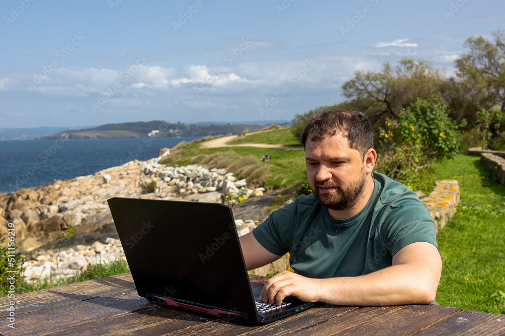 A man with a beard, pensive, sits at a laptop at a wooden table on the ocean. Freshness. Speculation. Thoughts. Plans.