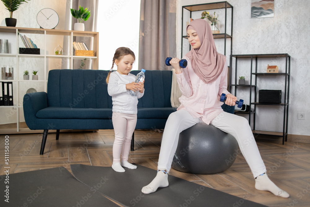 Obraz premium Happy Muslim family doing sports in morning at home. Beautiful Arab woman sitting on fitball and doing exercises with dumbbells while her little daughter sitting next to her with a bottle of water.