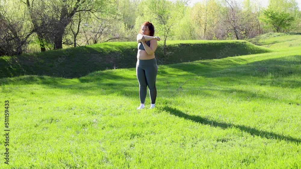 Young overweight woman in fitness suit stretch arm muscles standing on green lawn in park on sunny day, nature background. Healthy lifestyle, exercise and fitness outdoors. Sports activity concept. Young overweight woman in fitness suit stretch arm muscles standing on green lawn in park on sunny day, nature background. Healthy lifestyle, exercise and fitness outdoors. Sports activity concept.