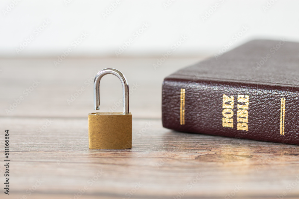Open lock (unlocked padlock) and closed Holy Bible Book on a wooden