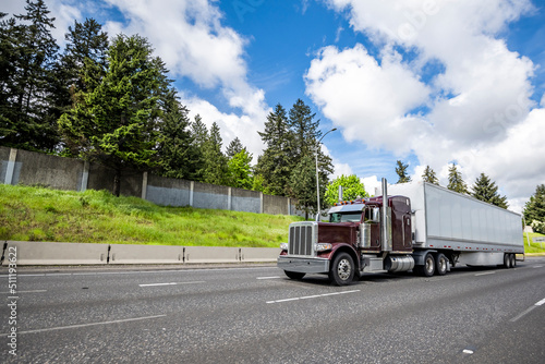 Classic burgundy big rig semi truck tractor transporting cargo in dry van semi trailer driving on the wide highway road with clouds sky