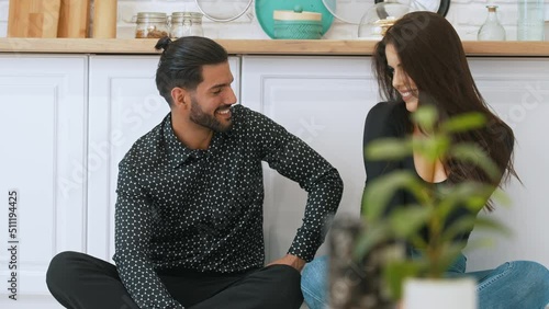 Cheerful millenial interracial married couple sitting barefoot on the floor with their legs crossed, smilig and talking together. High quality 4k footage photo