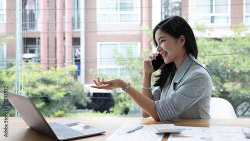 Attractive smiling young asian business woman relaxing at office, working on laptop computer, talking on mobile phone