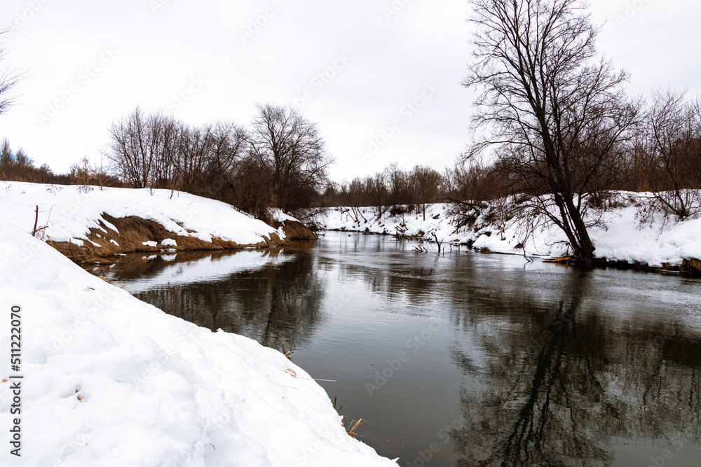 forest river with fast flow and small waves flows between snowy shores with trees and bushes late winter and early spring forest river with fast flow and small waves flows between snowy shores with trees and bushes late winter and early spring
