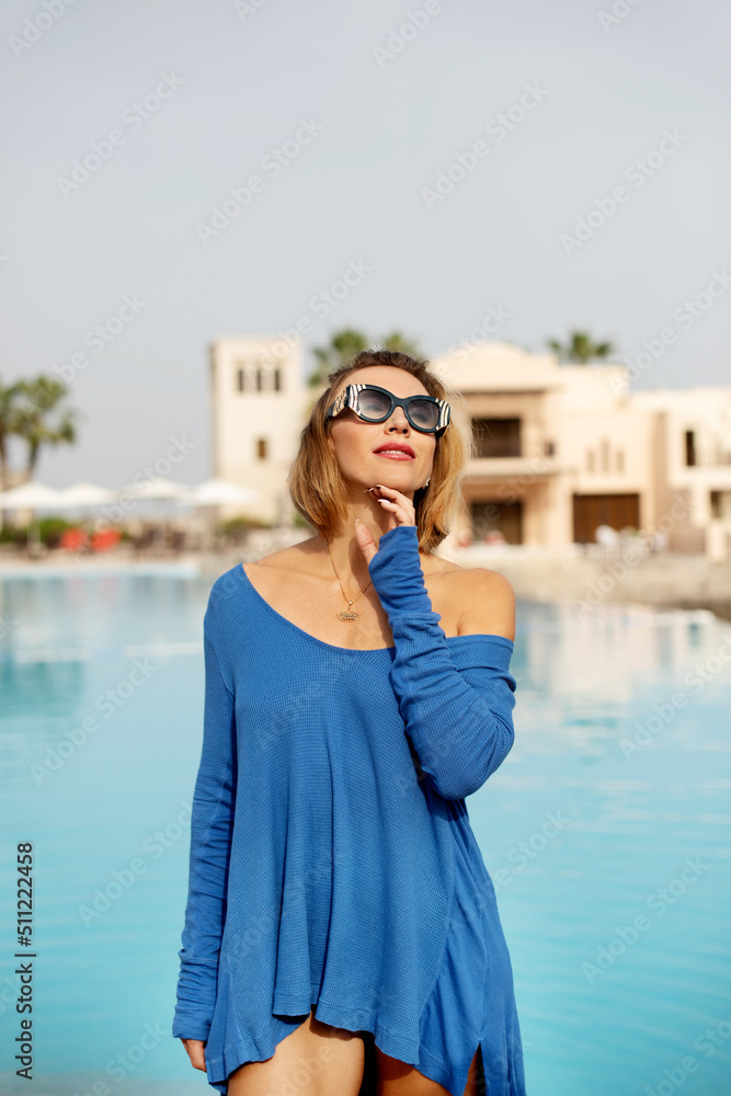 Fashionable summer portrait of a sexy tanned woman in a blue tunic posing by the pool in a luxurious tropical hotel