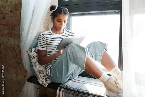 African girl sitting on windowsill and reading book online using digital tablet photo