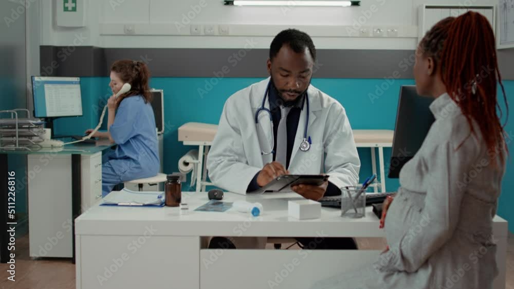 General practitioner taking notes on digital tablet at maternity checkup with pregnant patient. Male physician having conversation about pregnancy with future mother in medical office. General practitioner taking notes on digital tablet at maternity checkup with pregnant patient. Male physician having conversation about pregnancy with future mother in medical office.