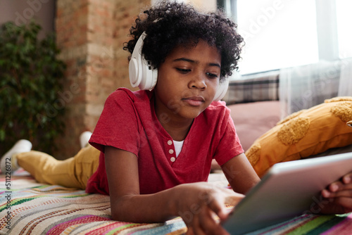 African little boy in wireless headphones lying on sofa and watching video online using digital tablet