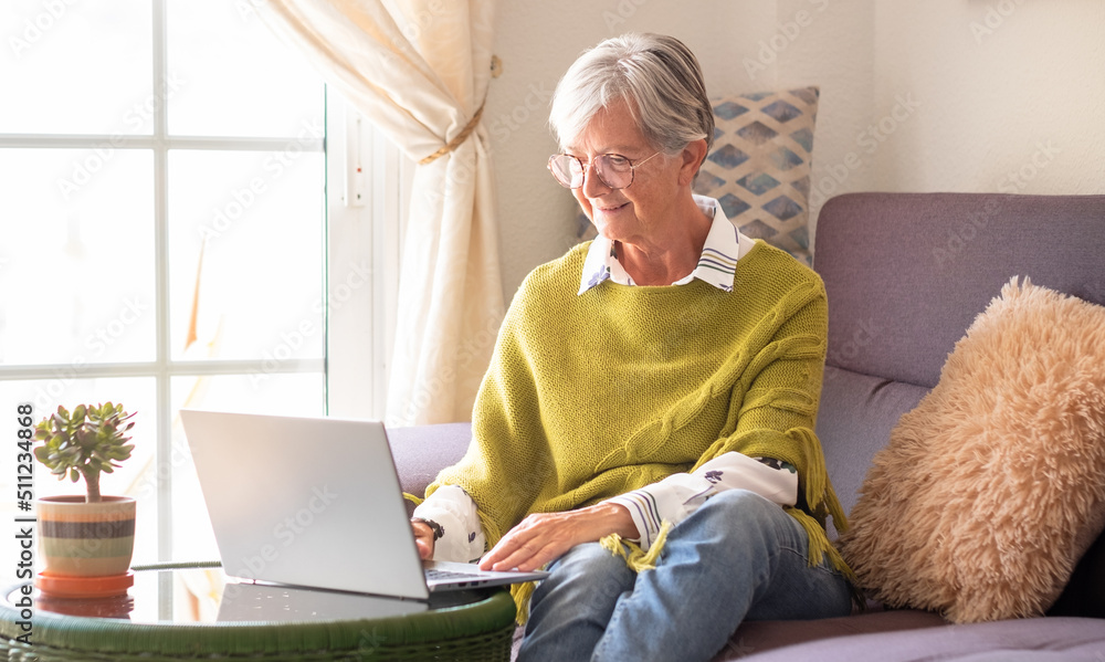 Smiling elderly woman working on laptop at home sitting on sofa in living room. Modern senior lady with green poncho browsing internet Smiling elderly woman working on laptop at home sitting on sofa in living room. Modern senior lady with green poncho browsing internet