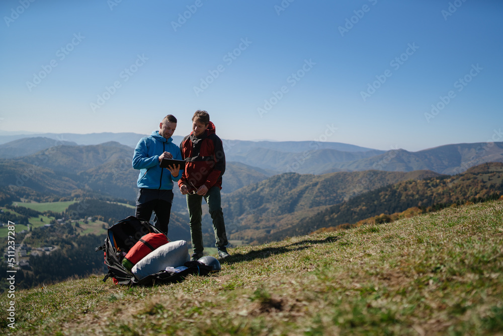Two paragliders men standing on the top of mountain with tablet and looking at view.