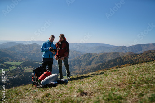 Two paragliders men standing on the top of mountain with tablet and looking at view.