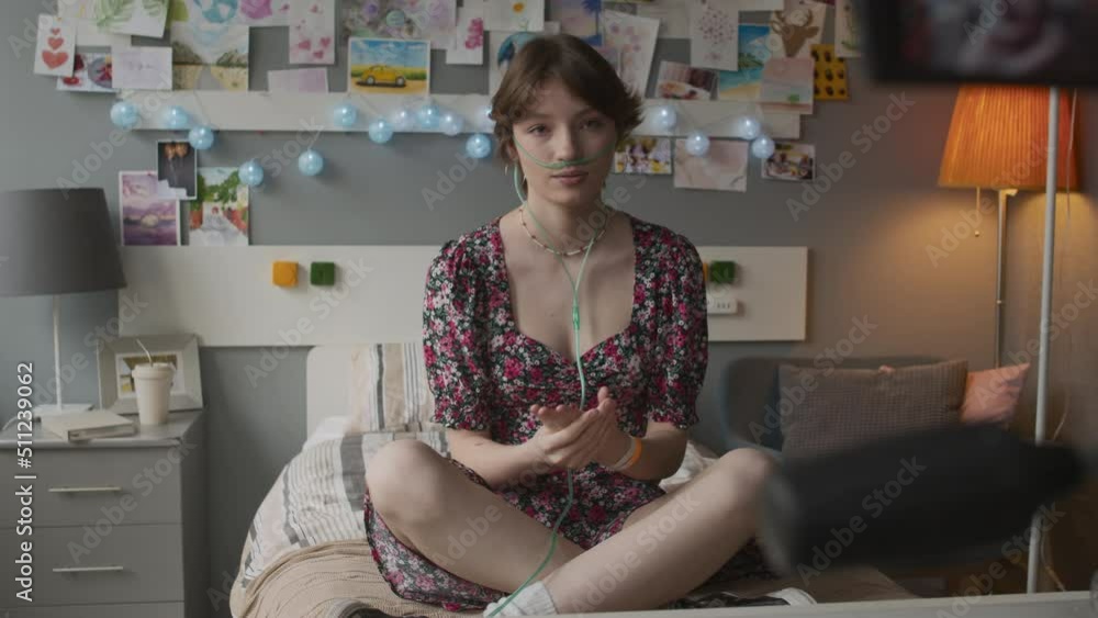 Teenage Caucasian girl with nasal cannula sitting on bed in decorated hospital ward, smiling, talking, filming vlog on video camera Teenage Caucasian girl with nasal cannula sitting on bed in decorated hospital ward, smiling, talking, filming vlog on video camera
