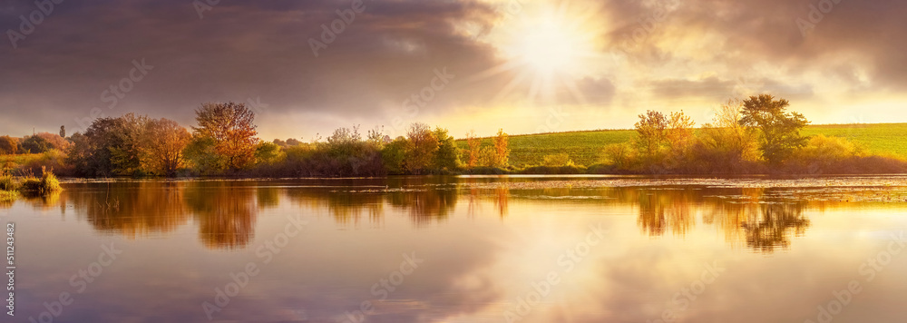 Autumn landscape with trees by the river and picturesque dark clouds reflected in the river water at sunset. The sun peeks through the dark clouds - obrazy, fototapety, plakaty 