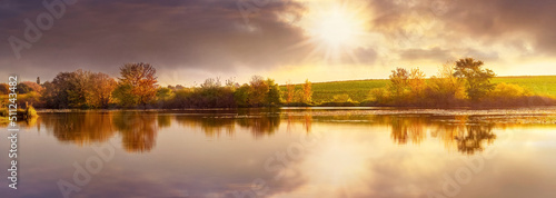 Autumn landscape with trees by the river and picturesque dark clouds reflected in the river water at sunset. The sun peeks through the dark clouds