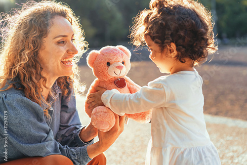 Curly single woman giving a teddy bear to her little daughter outdoors in the park - family lifestyle concept photo