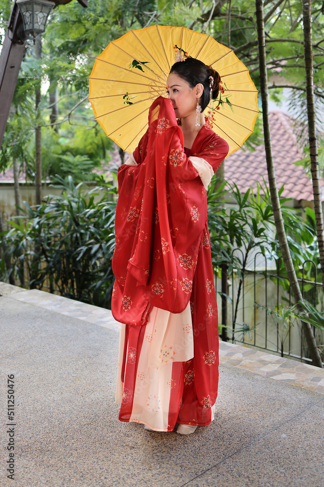 Fototapeta premium Young attractive Asian woman wearing tadeonal Chinese red hanfu long skirt dress costume scarf hairpin earing decorated umbrella outdoor green garden patio
