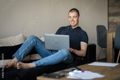 06.09.2022 - Pleven,Bulgaria - Enjoying time at home. Young smiling men working on laptop while laying in a big comfortable sofa at home. Job Seeking, work at home concept