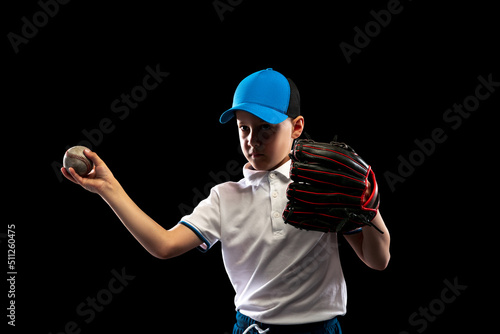 One kid, little baseball player in blue-white uniform leaning to play baseball isolated on black studio background. Concept of sport, achievements, studying, competition