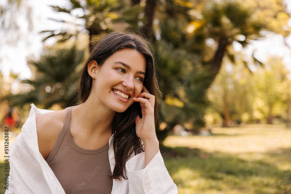 Cheerful young caucasian woman smiles with teeth enjoying nature on sunny day. Brunette with flowing hair wears top and shirt. Summer vacation concept Cheerful young caucasian woman smiles with teeth enjoying nature on sunny day. Brunette with flowing hair wears top and shirt. Summer vacation concept