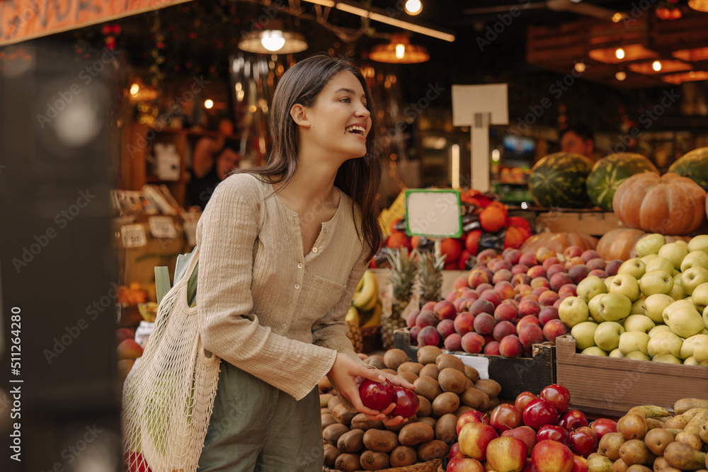 Happy caucasian young girl chooses fruits at food market in center of european city. Brunette wears casual clothes with shopping bag. Concept of natural organic products