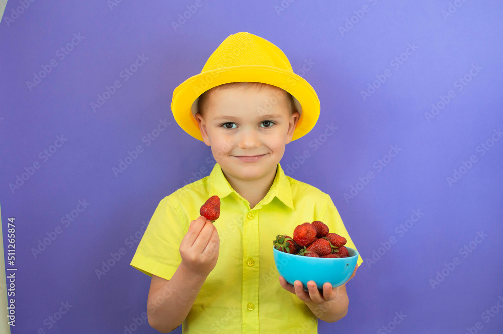 A happy smiling child boy in a summer yellow hat is eating strawberries on a purple background. The concept of healthy baby food. Happy summer childhood. Selective focus. A happy smiling child boy in a summer yellow hat is eating strawberries on a purple background. The concept of healthy baby food. Happy summer childhood. Selective focus.