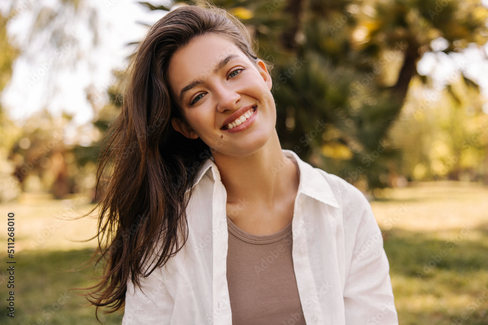 Smiling young caucasian girl looking at camera on blurred nature background during day. Brunette wears casual clothes in spring. People emotions, lifestyle and fashion concept.
