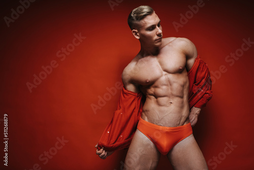 Sexy young man in red speedo and jean jacket stripping at red background. Handsome shirtless guy taking off his red jacket. Fitness male model in studio. Muscular sportsman with six pack abs. photo