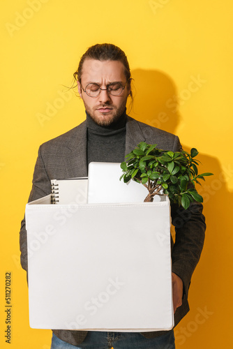 Upset man holding box with personal items after job resignation against yellow background