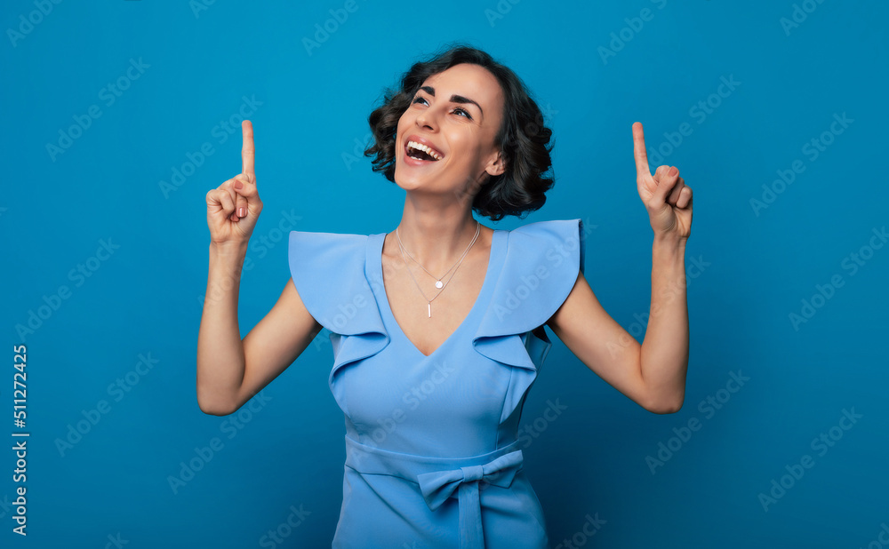 Close-up photo of a happy beautiful young woman in a blue dress, who is pointing with her index finger and isolated on blue background Close-up photo of a happy beautiful young woman in a blue dress, who is pointing with her index finger and isolated on blue background