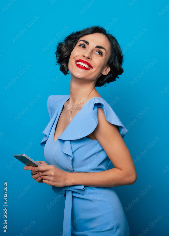 Online. Apps. Typing. Communication. Internet. User. Close up portrait of smiling cute stylish brunette woman with modern smart phone in hands isolated on blue background Online. Apps. Typing. Communication. Internet. User. Close up portrait of smiling cute stylish brunette woman with modern smart phone in hands isolated on blue background