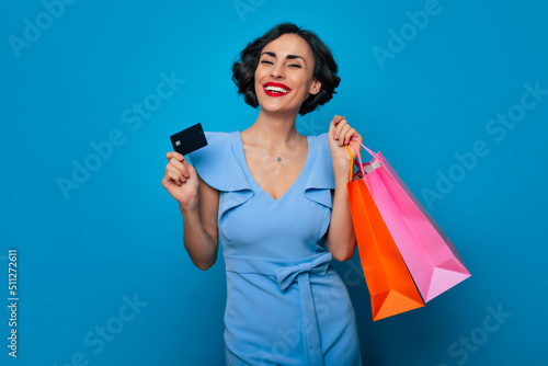 Close up portrait of smiling beautiful stylish brunette woman with shopping bags and credit card in hands isolated on blue background