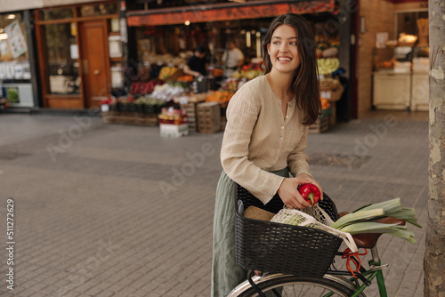 Attractive young caucasian girl looking away packs food into bicycle basket. Brunette wears casual clothes on street. Mood, lifestyle, concept. photo