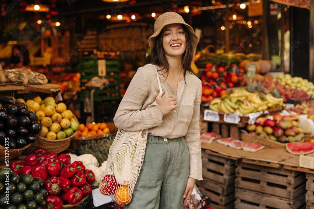 Nice caucasian young woman with shopping bag smiling looks at camera standing in vegetable row of market. Brunette wearing casual clothes is shopping on weekends. Lifestyle concept Nice caucasian young woman with shopping bag smiling looks at camera standing in vegetable row of market. Brunette wearing casual clothes is shopping on weekends. Lifestyle concept