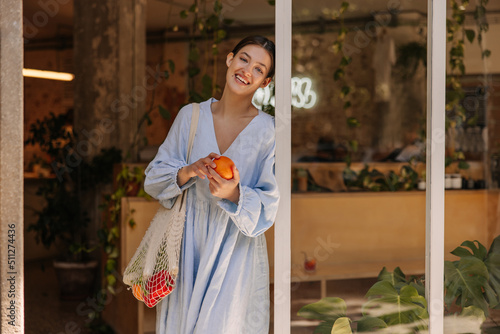 Positive young caucasian woman with shopping bag smiling looking at camera, holding orange. Brunette wears blue sundress on casual day. People sincere emotions lifestyle concept.