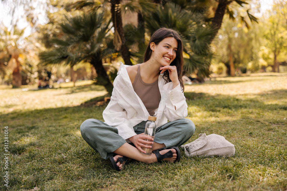 Happy young caucasian woman spends time outdoors sitting on grass. Brunette with beaming smile wears casual clothes. City life concept Happy young caucasian woman spends time outdoors sitting on grass. Brunette with beaming smile wears casual clothes. City life concept