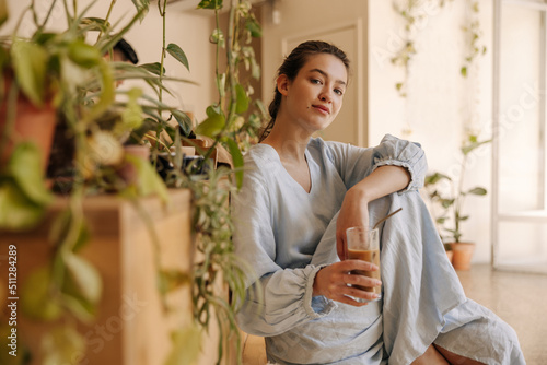 Pretty young caucasian woman with coffee smoothie enjoys pastime sitting indoors. Brunette wears blue sundress on summer day. Relaxation concept photo