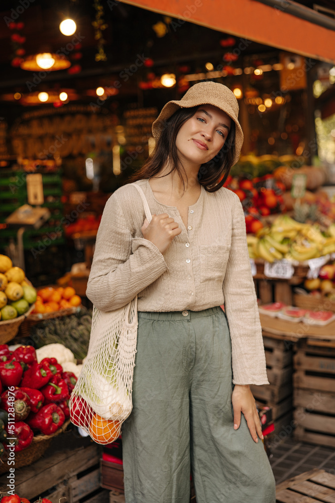Cute young caucasian woman spends leisure time at market with vegetables and fruits outdoors. Brunette wears casual clothes, hat and bag. Shopping concept