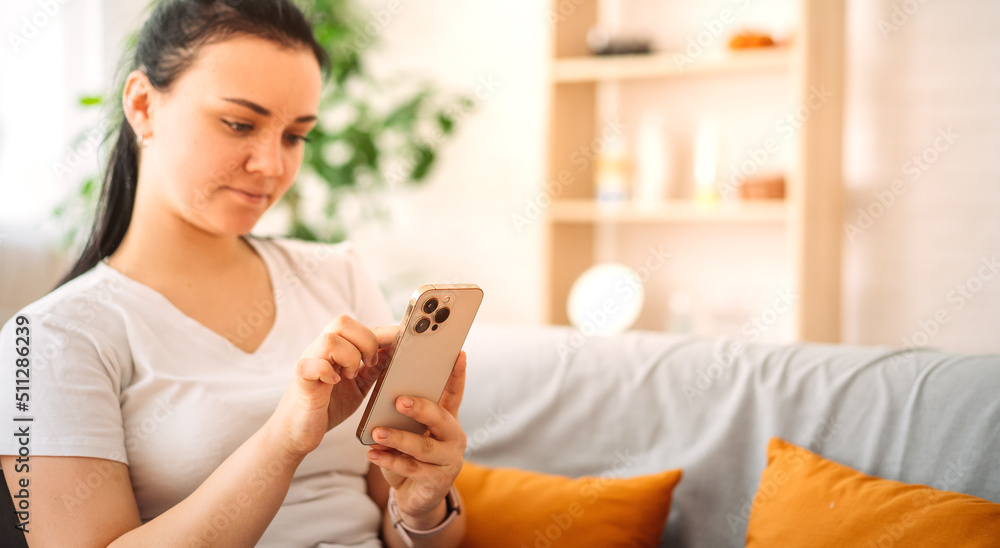 a brunette girl checks social networks at home. A smiling young girl of European appearance, using a phone, playing games, shopping online, ordering delivery on the couch. a brunette girl checks social networks at home. A smiling young girl of European appearance, using a phone, playing games, shopping online, ordering delivery on the couch.