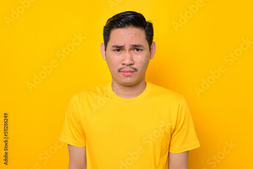 Dazed young Asian man in casual t-shirt looking at camera and emotionally reacting to news isolated on yellow background. People lifestyle concept