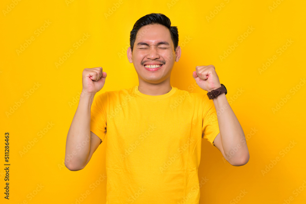 Overjoyed young Asian man in casual t-shirt celebrating big luck and success with raised fists isolated on yellow background. People lifestyle concept Overjoyed young Asian man in casual t-shirt celebrating big luck and success with raised fists isolated on yellow background. People lifestyle concept