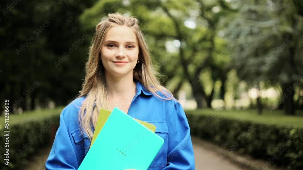 Portrait of student girl walking down with books near the park and looking at the camera. Portrait of attractive blond girl. Portrait of student girl walking down with books near the park and looking at the camera. Portrait of attractive blond girl.