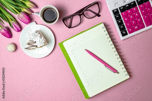Flat lay view of desktop surface, tulips, coffee with cake and notepad with calculator on pink light background, holiday in financial sphere