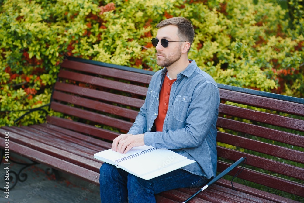 Fototapeta premium Blind man reading braille book, sitting on bench in summer park, resting