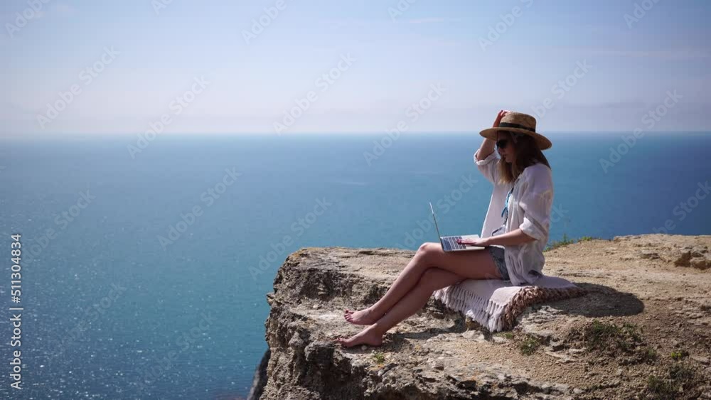 Successful business woman in yellow hat working on laptop by the sea. Pretty lady typing on computer at summer day outdoors. Freelance, digital nomad, travel and holidays concept. Successful business woman in yellow hat working on laptop by the sea. Pretty lady typing on computer at summer day outdoors. Freelance, digital nomad, travel and holidays concept.