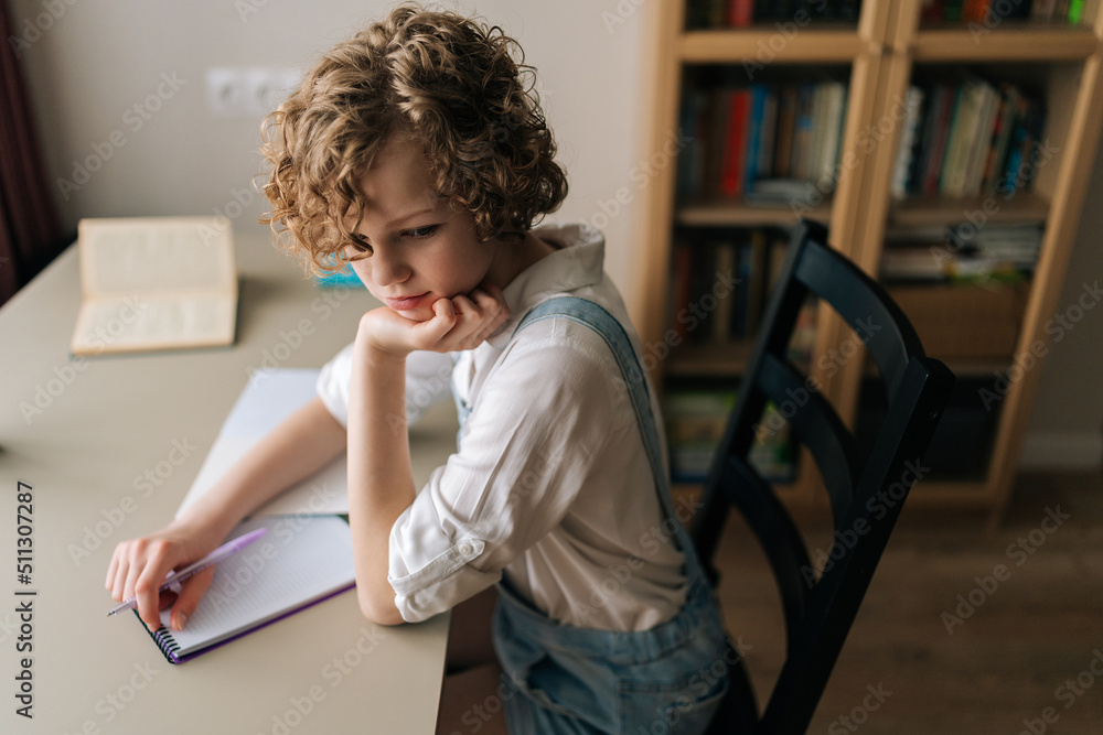 High-angle view of thoughtful little child school girl sitting at desk near window and thinking with pen in hands looking away. Pretty pensive schoolgirl with curly hair sitting at wooden table. High-angle view of thoughtful little child school girl sitting at desk near window and thinking with pen in hands looking away. Pretty pensive schoolgirl with curly hair sitting at wooden table.