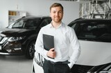 portrait of open-minded professional salesman in cars showroom, caucasian man in white formal shirt stands next to luxurious car and looks at camera