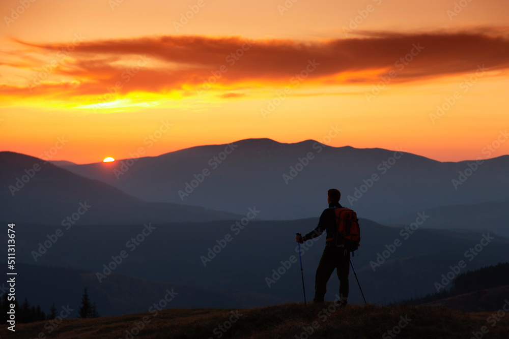A man with a backpack is outdoor at sunset. Orange sky. Silhouettes of mountains peaks